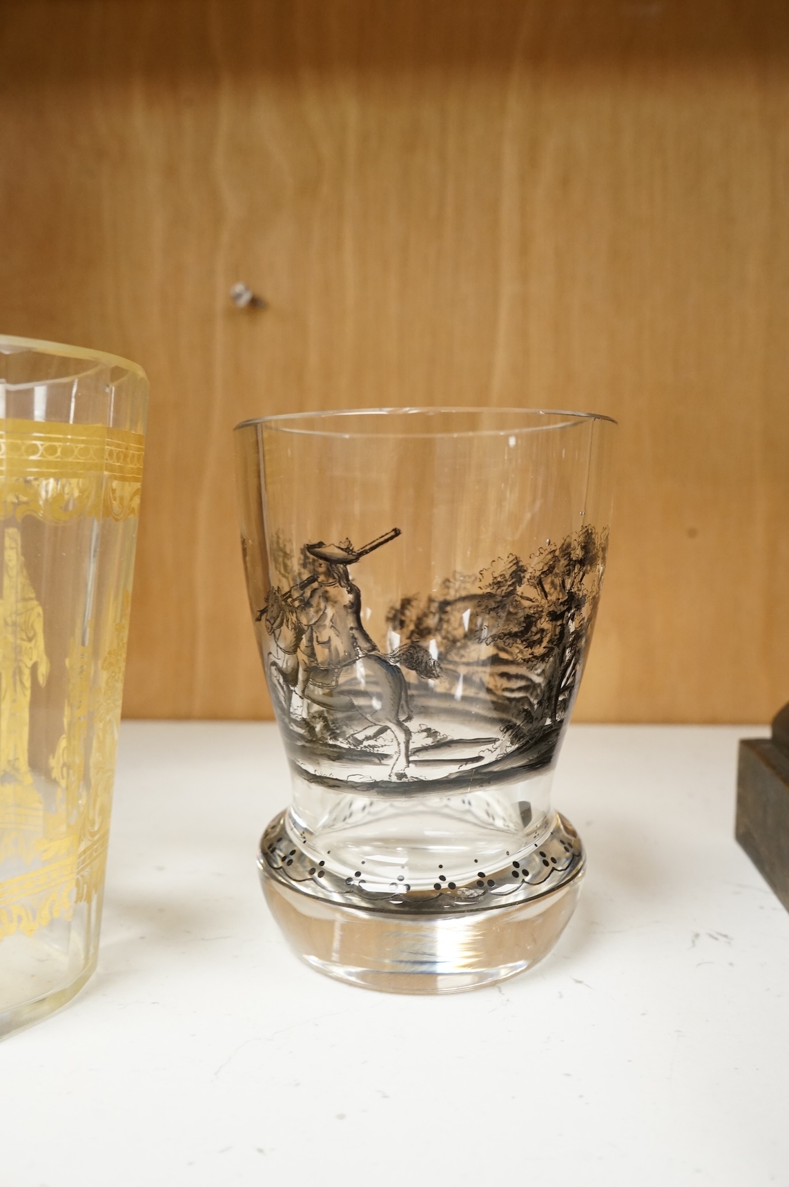 An Italian gilt-decorated and faceted beaker, inscribed Daniel Delvinis, the reverse with papal coat of arms, and a German Schwarzlot black enamelled ‘hunting’ beaker, largest 12cm (2)